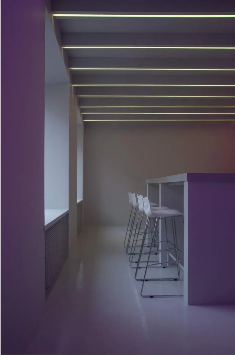 Modern corridor with purple walls, linear ceiling lights, and bar seating area with white chairs along a counter