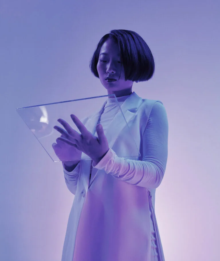 Woman with short dark hair holding transparent glass tablet, wearing white long-sleeved top against purple-toned background