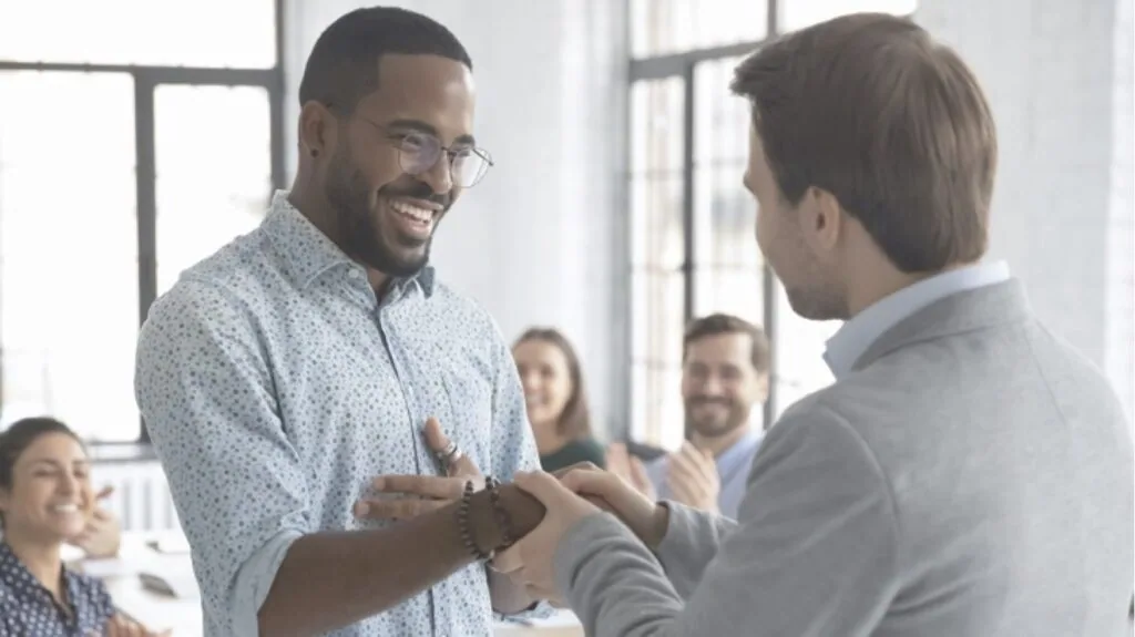 Two men shaking hands in an office while colleagues watch in background