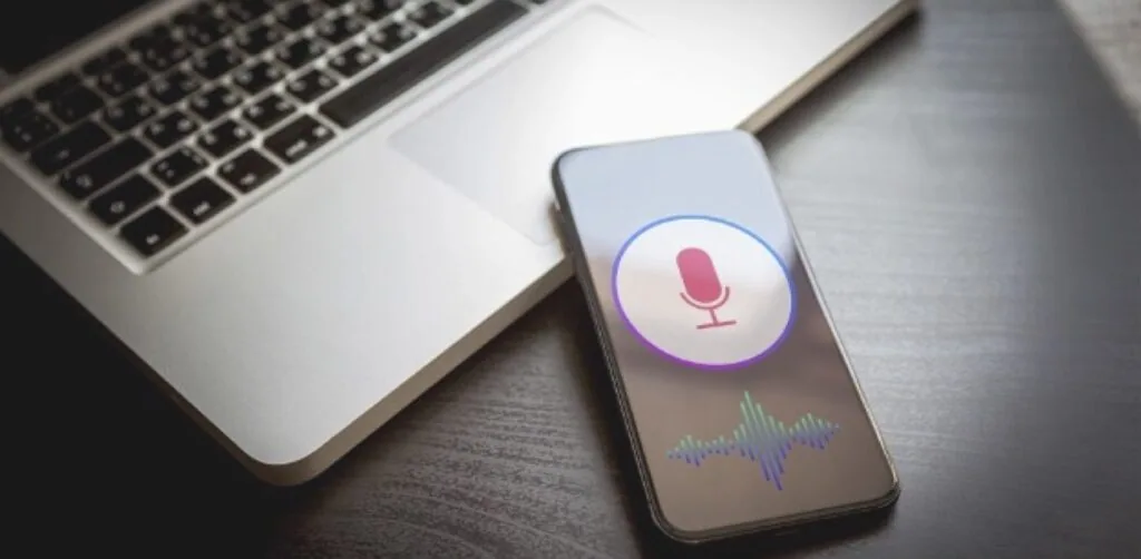 Smartphone displaying voice recognition interface with microphone icon and audio waveform next to laptop on desk