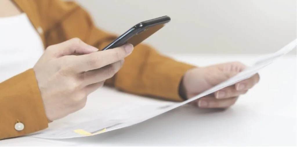 Person in mustard-colored sleeve holding smartphone while reviewing document on white surface